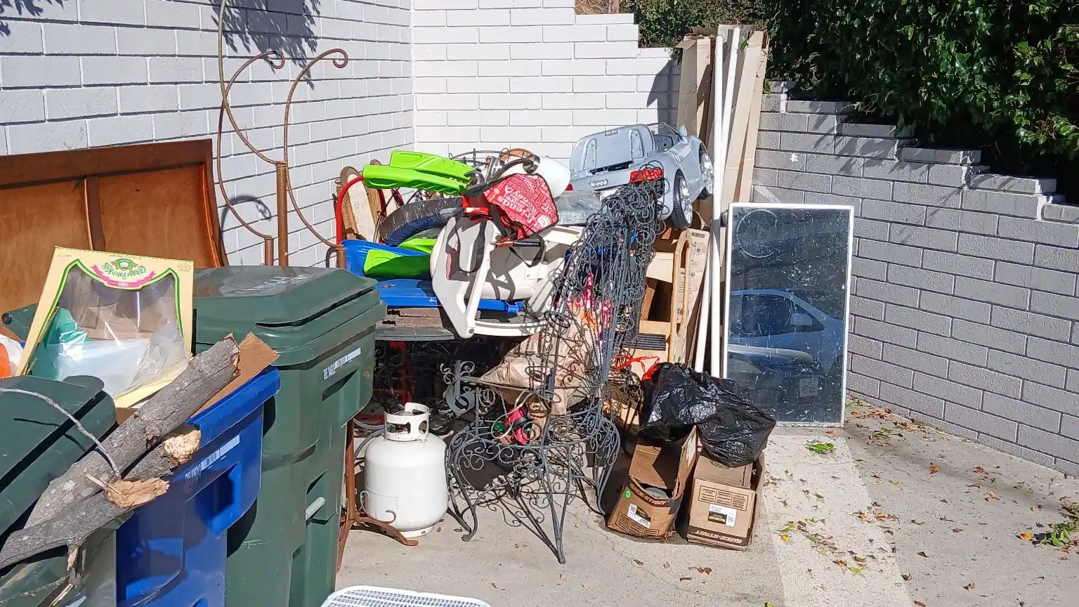 Stored household items, chairs, bins and boxes stacked along a side yard wall for pickup