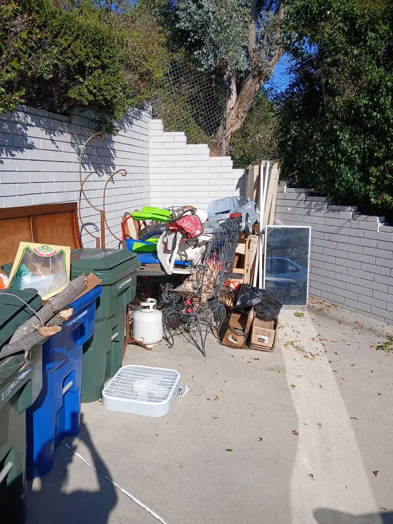 Stored household items, chairs, bins and boxes stacked along a side yard wall for pickup