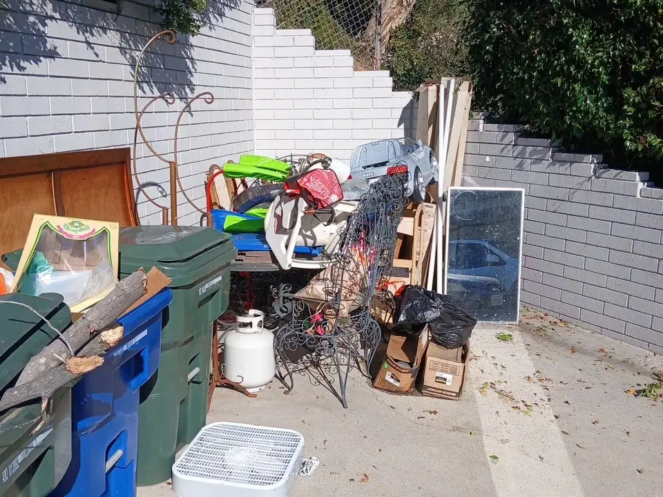 A side yard by a white brick wall piled with furniture; bins; boxes and a propane tank